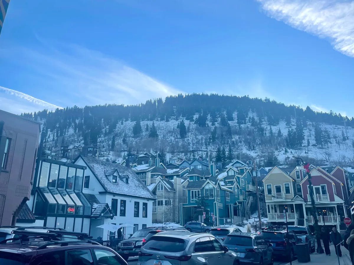 A bottom-up view of colorful buildings and parked cars in Park City with a tree filled snowy mountain in the towering distance. 