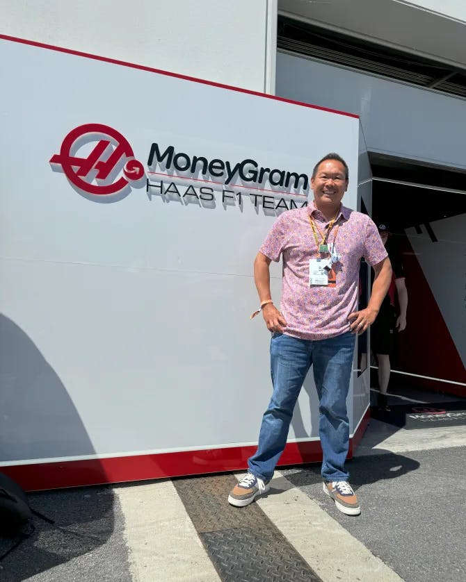 Robert in a pink shirt and jeans standing in front of a white wall with a sign reading “MONEYGRAM” outdoors on a sunny day