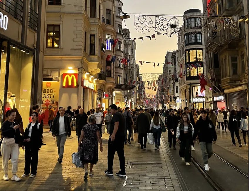 An assortment of people walking down a stone street with old architecture, a McDonalds logo, and decorative arches and flags in the surrounding areas. 