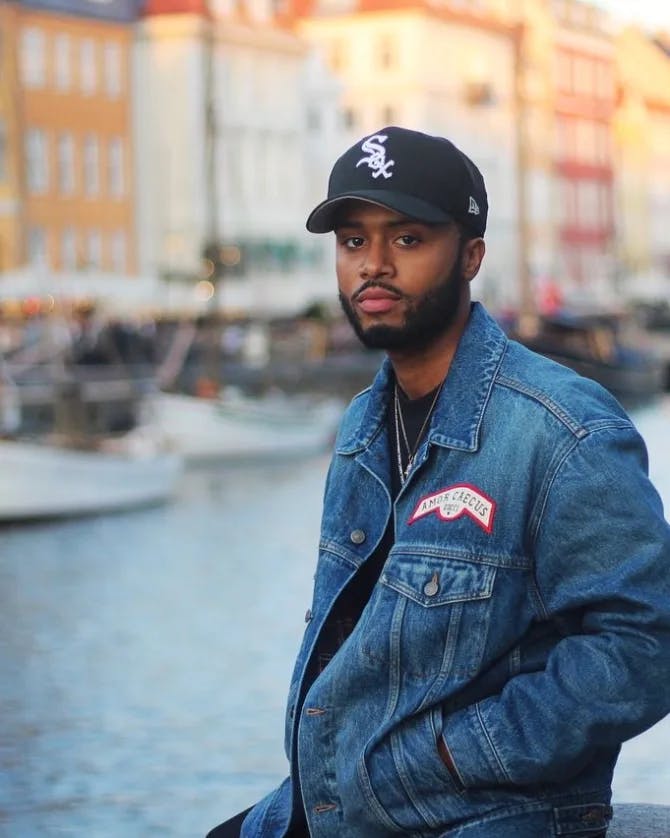 Mark wearing a denim jacket and a baseball cap posing for a portrait photo along a canal with colorful buildings in the background