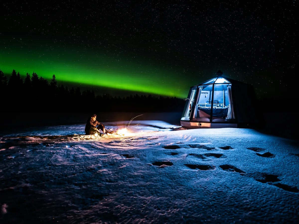 A serene night scene with the mesmerizing northern lights, a person sitting outside on the snowy ground near a makeshift camping portal and a cozy campfire.