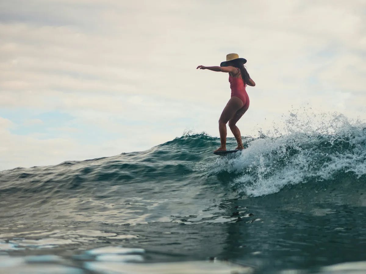 A surfer in an orange swimsuit and straw hat riding a wave, set against an overcast sky.