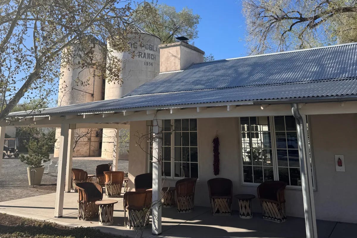 An image of an inn with white paned windows, an outdoor patio with chairs and tables, factory style architecture in the background and trees in the surrounding area. 