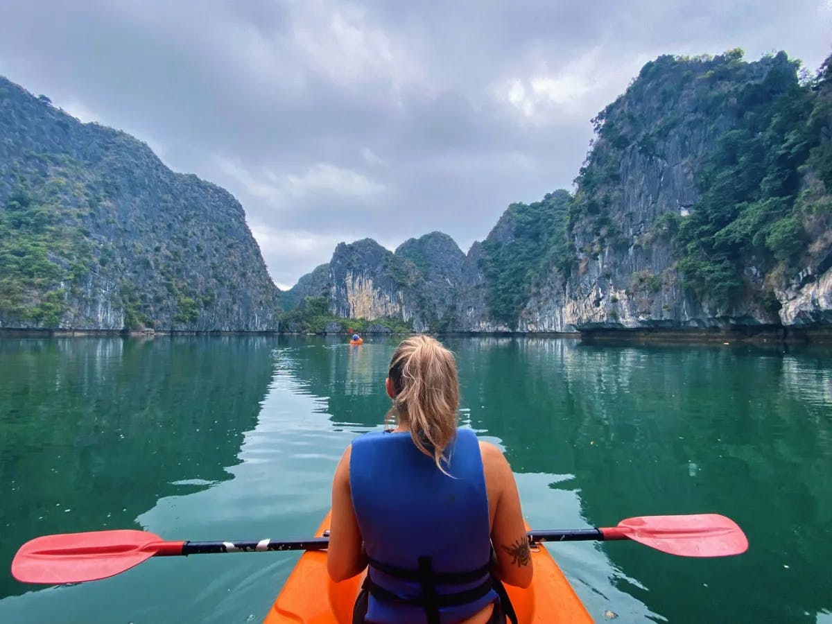 A person is kayaking in a serene body of water surrounded by towering, lush green limestone cliffs under a cloudy sky.