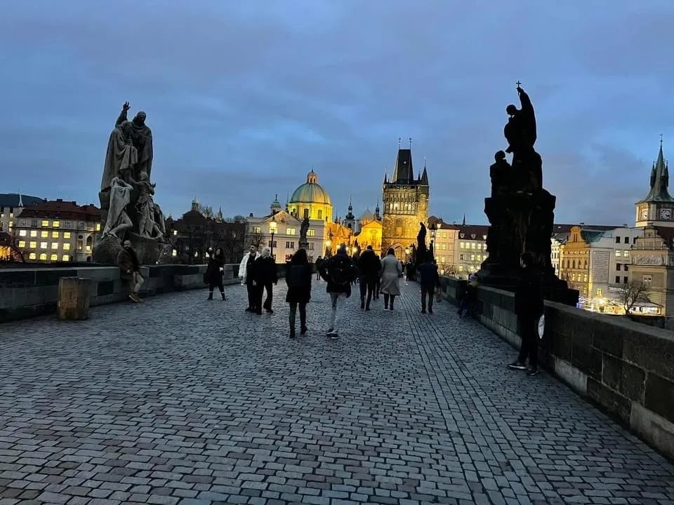 A stone bridge with several pedestrians during twilight, with historical statues and the Old Town bridge tower