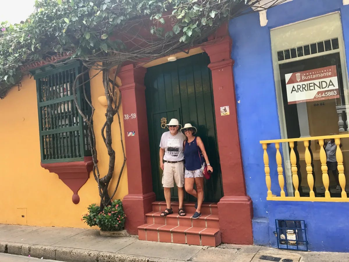 A vibrant snapshot of urban life: two individuals pose by a colorful building adorned with greenery and a sign for “Bustamante ARRIENDA.”