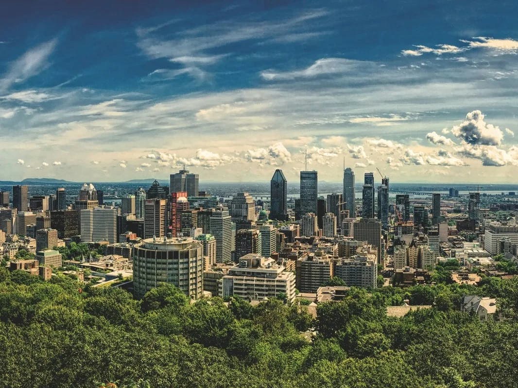 An aerial view of the Montreal skyline during the day, with buildings behind trees and clouds in a blue sky.