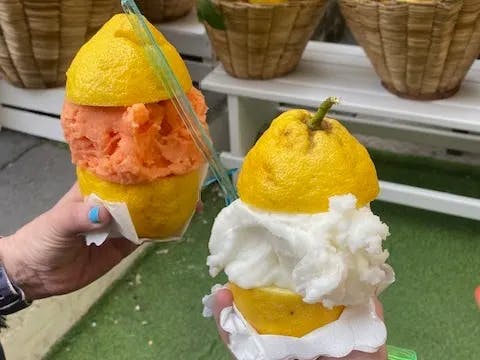 The image shows a close-up of two hands holding halves of two different citrus fruits filled with sorbet, against a background featuring more citrus fruits and baskets.