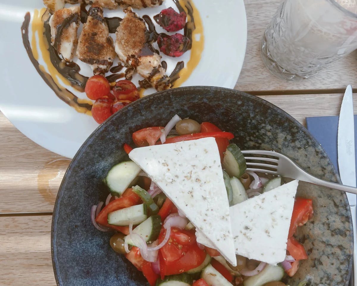 Greek salad with pita on a black plate and meat on white plate on a brown table.