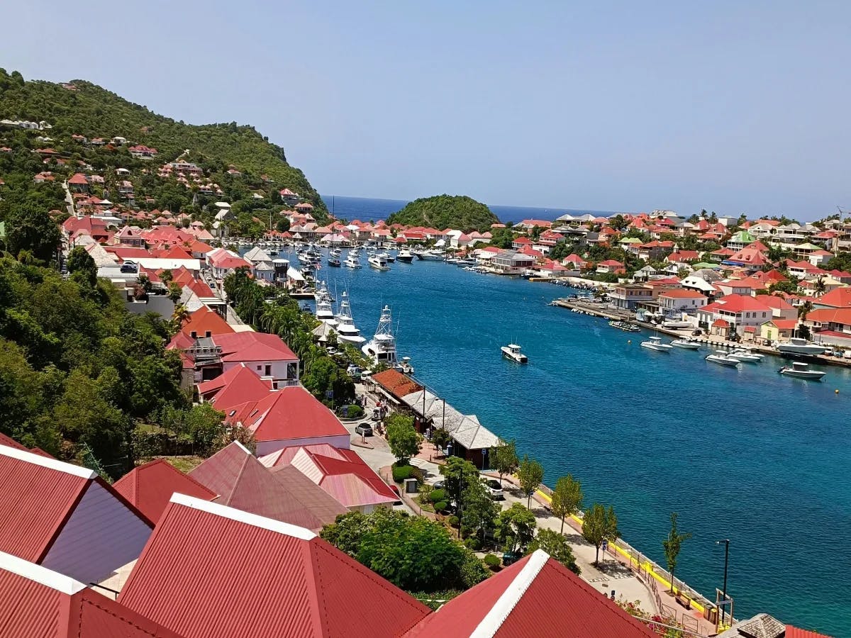 A picturesque coastal town with red-roofed buildings and a boat-filled harbor, nestled against green hills.