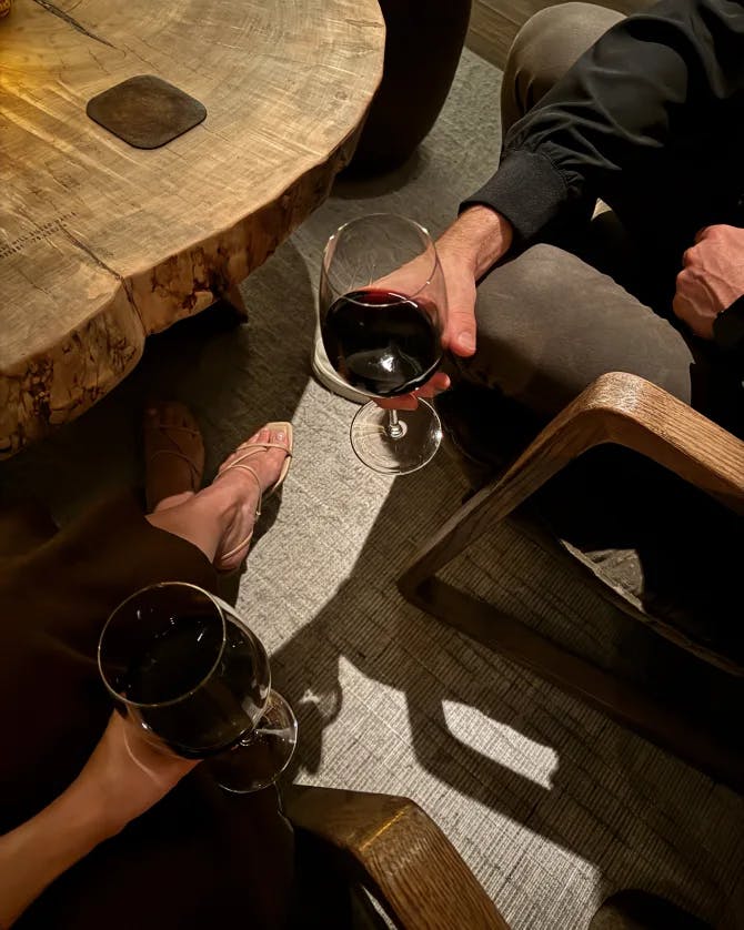 View of two people each holding a glass of red wine seated in wooden chairs by a wooden table 
