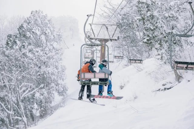 Two skiiers on a chair lift over a snowy mountain during winter, one of the beautiful Japan seasons.