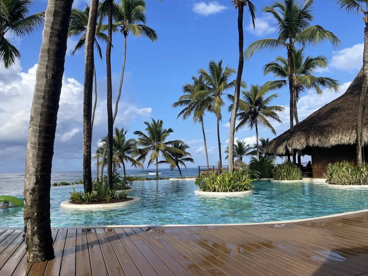 A picture of the resort's pool area with small islands of greenery in the middle of the pool, plus a large palapa-style building to the right and many palm trees around the area