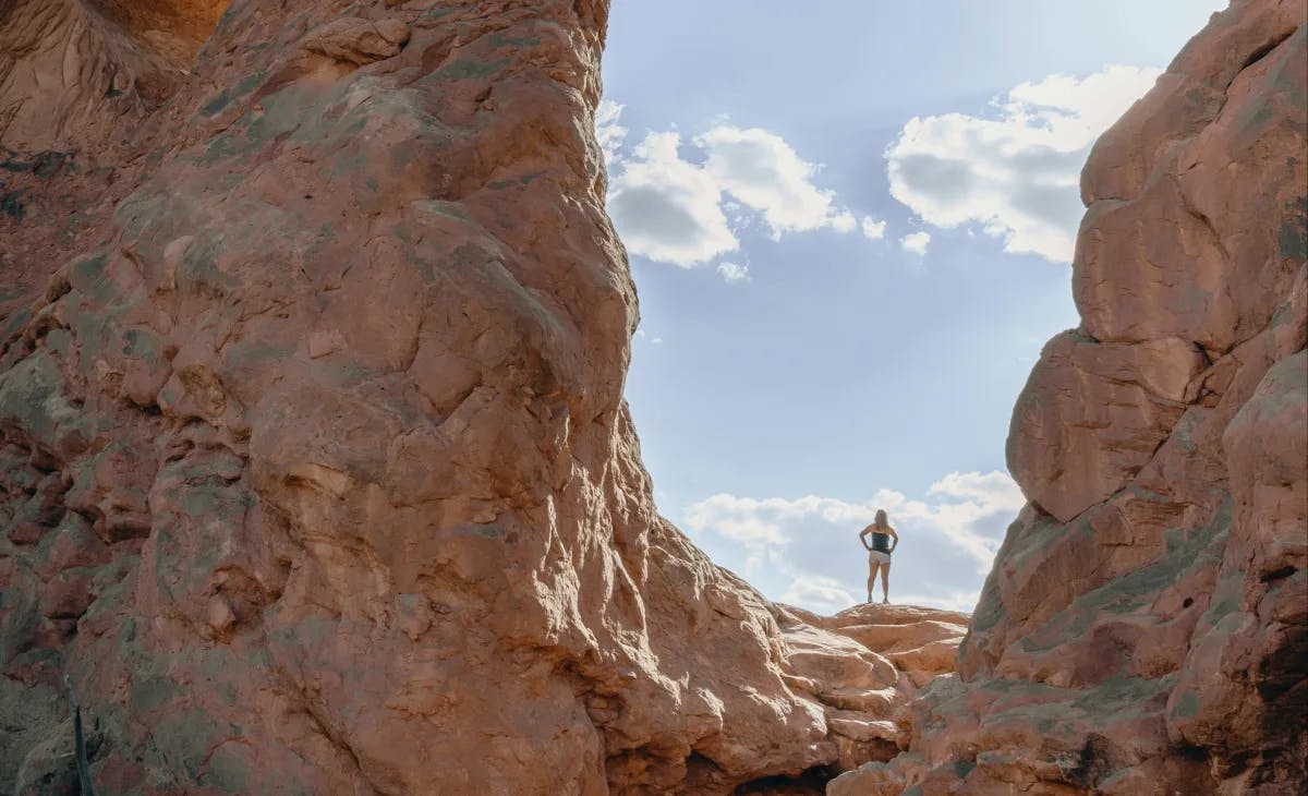 A person standing between two large red rock formations looking out at a blue partly cloudy sky.