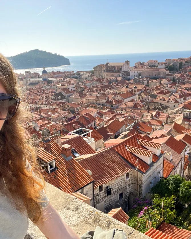 Kayla in sunglasses taking a selfie from a viewpoint overlooking orange rooftops in a coastal town, the ocean visible behind her