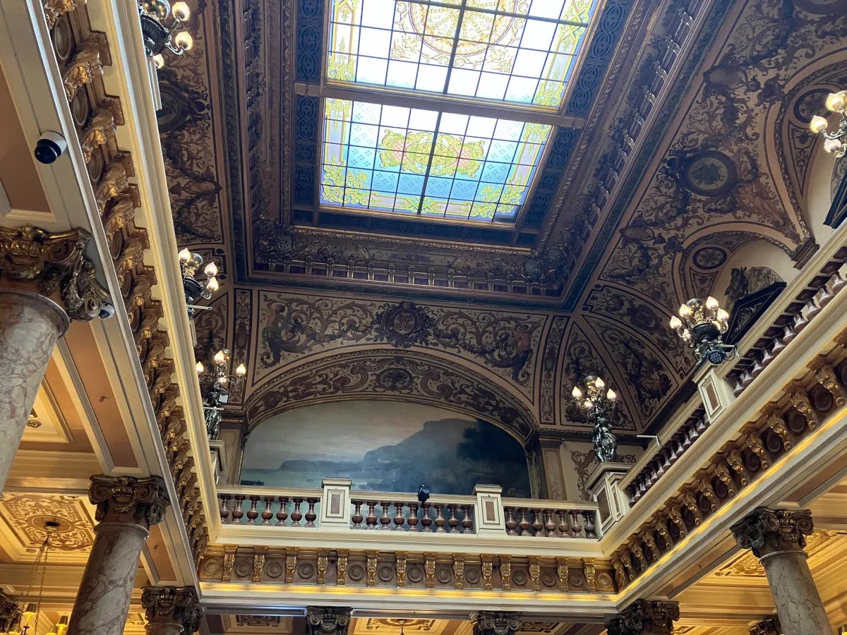 The image showcases an ornate ceiling with elaborate moldings, chandeliers, and a stained glass skylight, reflecting grandeur and elegance.