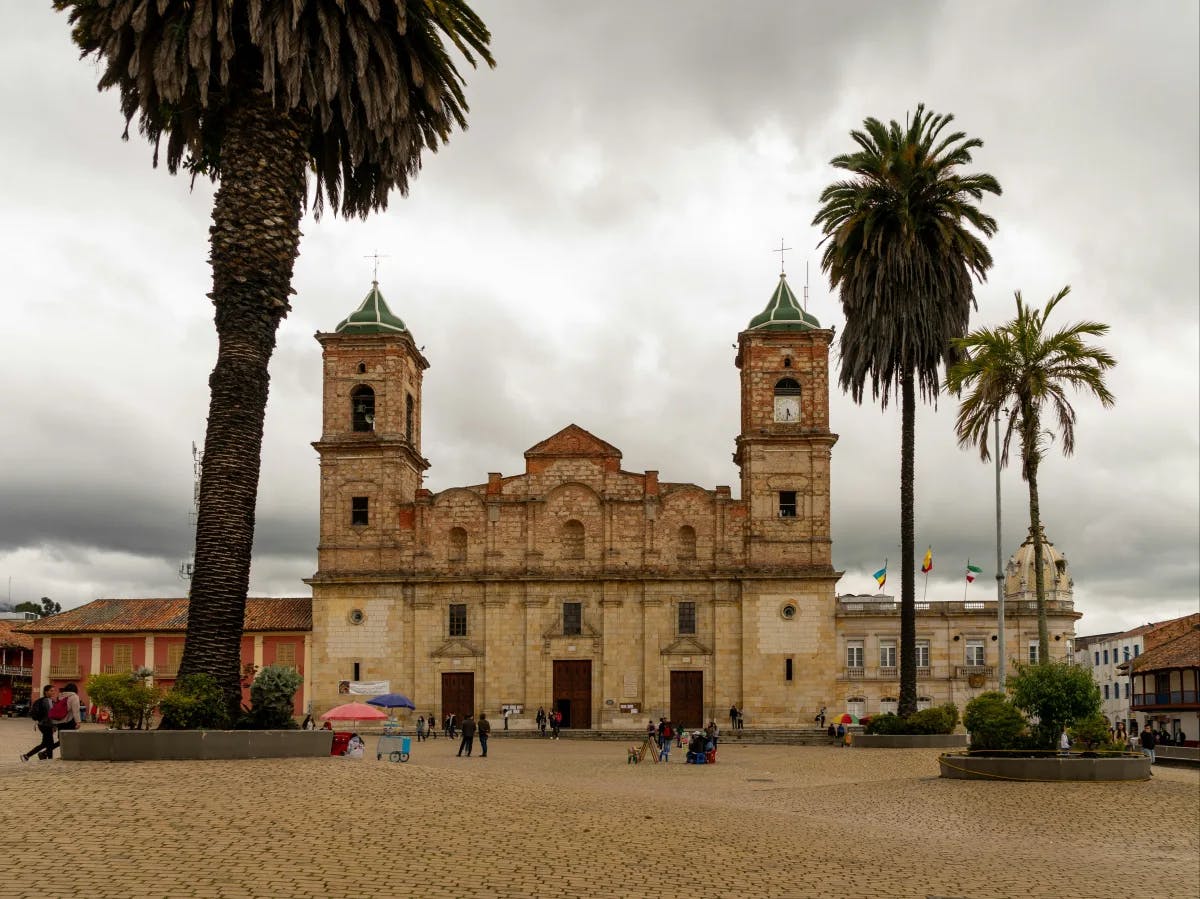 A historic stone edifice with twin bell towers and a central dome, flanked by palms and bustling with square activity under a cloudy sky.