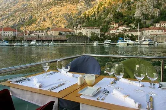 A wooden table with white tablecloths and wine glasses on a riverfront patio with foliage-covered hills in the background.