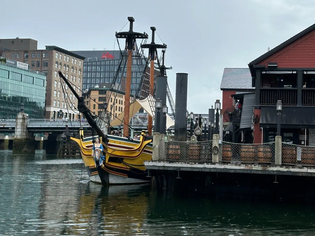 A large, ornate ship with multiple masts is docked near urban buildings, reflecting on the water's surface.