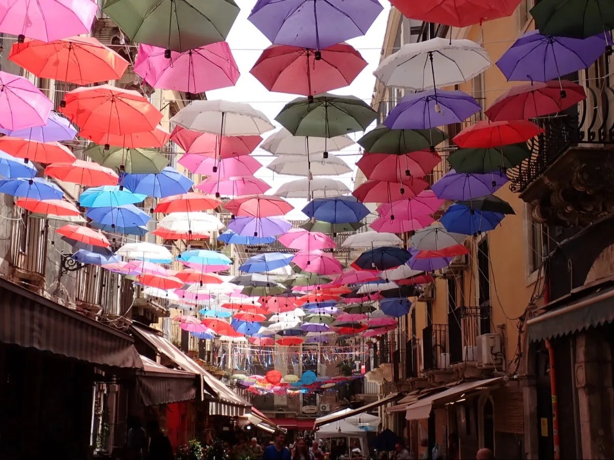 The image shows a vibrant display of numerous colorful umbrellas suspended above a narrow street, creating a whimsical canopy against the sky.