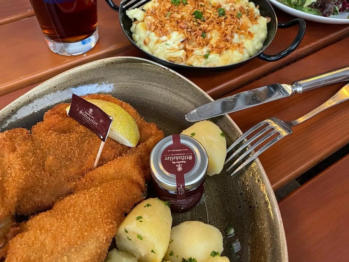 Two platters of food with silverware resting on one of them and a beverage in the top left corner, all positioned on top of a wooden table. 