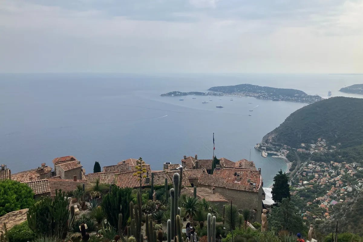 A picturesque coastal vista, showcasing terracotta rooftops amidst lush greenery, with a tranquil sea and boats under a soft, hazy sky.