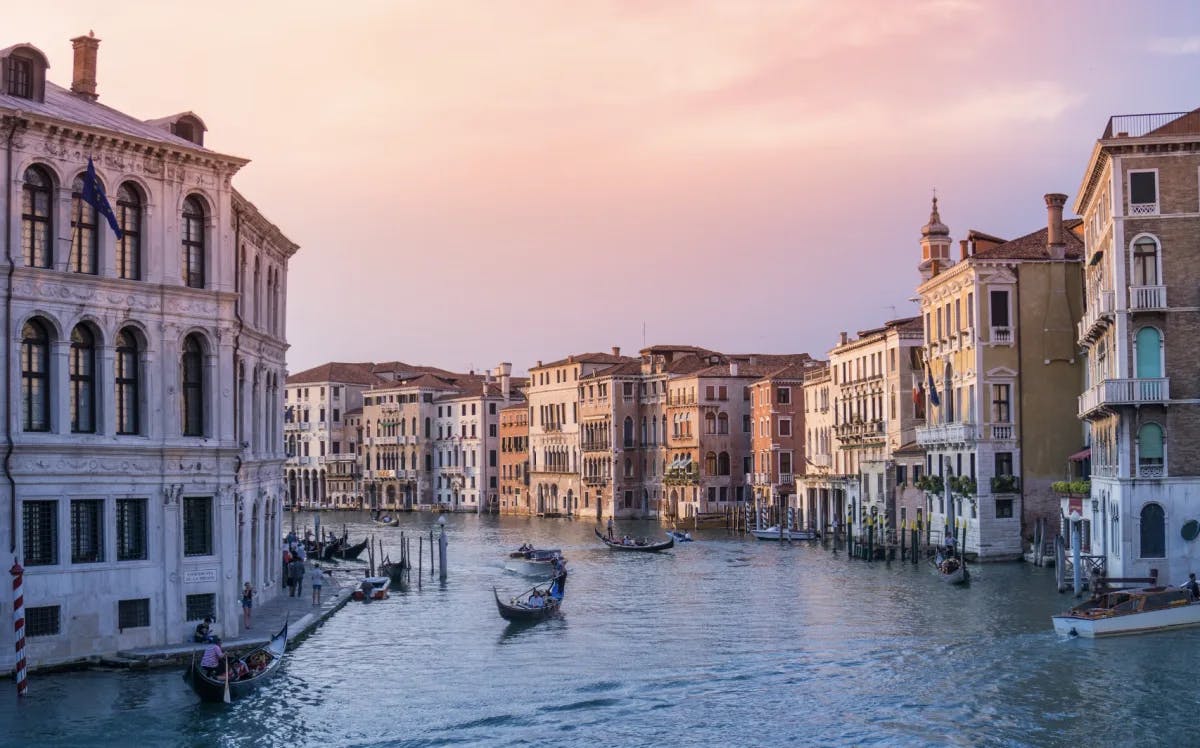 A view of a canal with gondolas floating down and beautiful buildings complete with old Venetian architecture and a pink and purple sunset in the background. 
