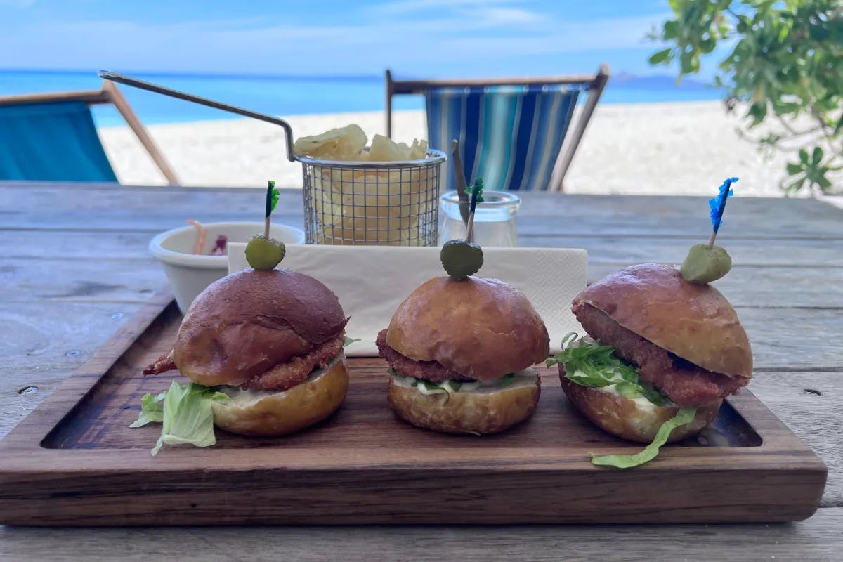 A picture of three burgers on a table with the beach at the background.