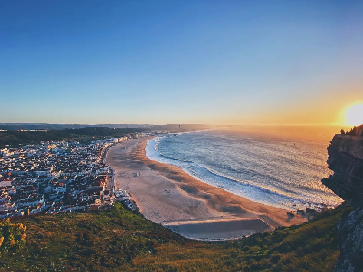 The image depicts a coastal town at sunset with a wide beach and the sun casting warm light over the scene.