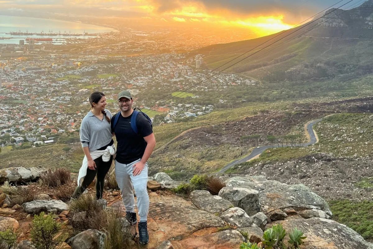 Two individuals stand atop a rocky elevation, gazing over a city illuminated by the warm glow of sunset.