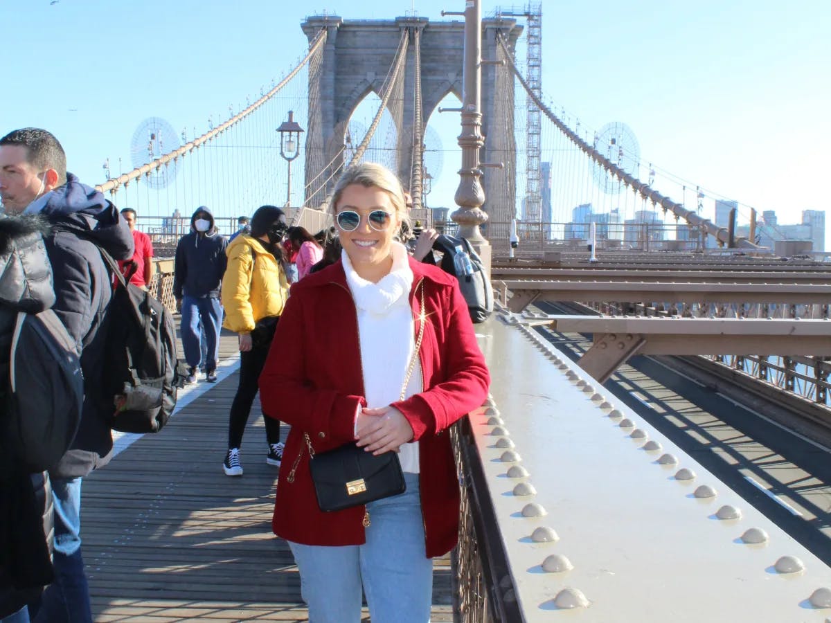 A woman wearing a white top and red jacket posing on the Brooklyn Bridge with many people walking around in the surrounding areas on a bright, sunny day. 