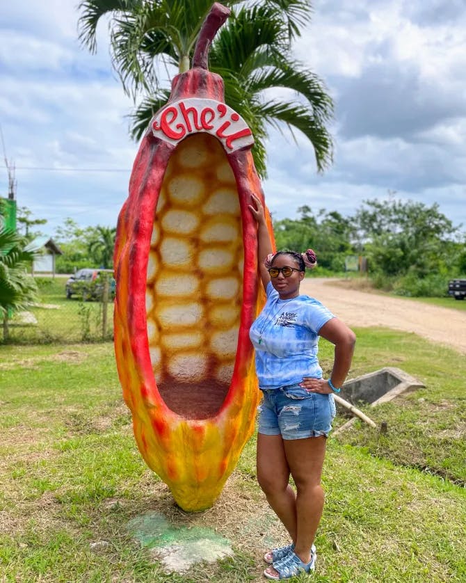 Jia wearing a blue top and denim shorts posting with a corn statue outside with palm trees and grass 