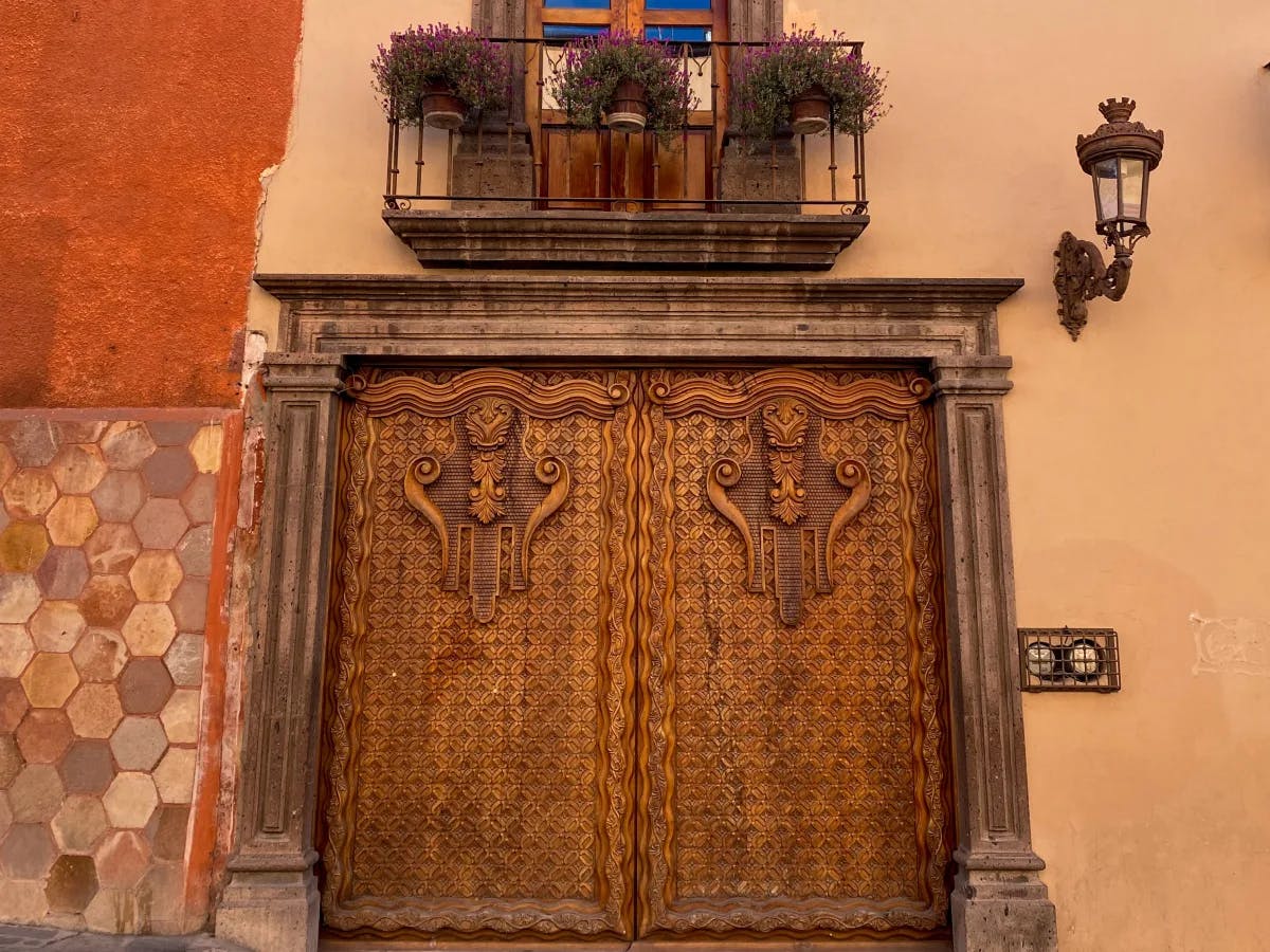 A wooden double doorway with intricate detailing, stone work, a lantern sconce and metal balcony with potted flowers above it.