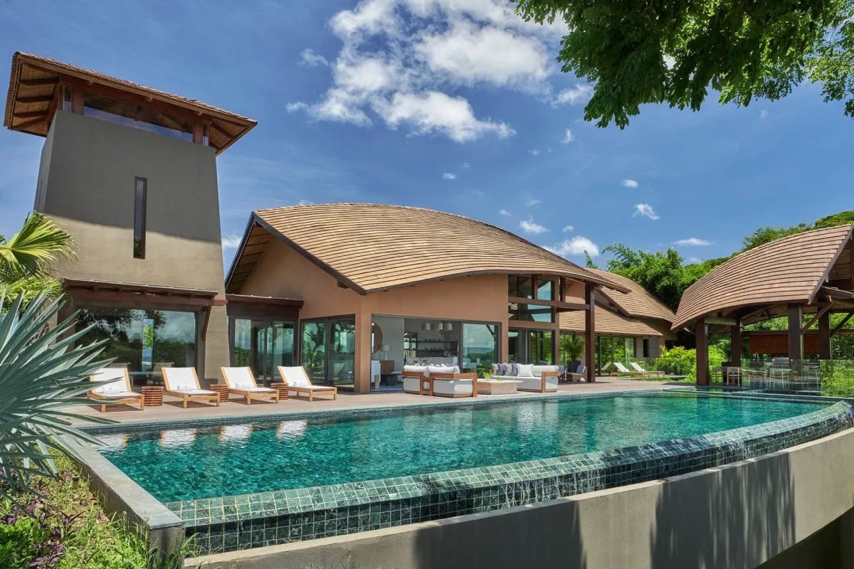 A lengthy turquoise-blue pool fills the foreground while jungle trees fill the border and a contemporary villa with loungers sits in the background