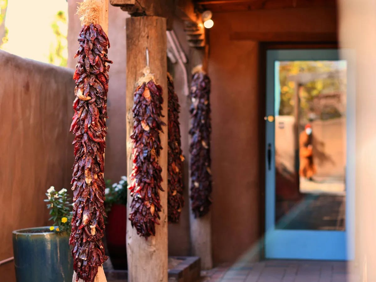 The image displays a row of dried red chili pepper ristras hanging from wooden beams in a rustic setting, with a blurred figure walking past a glass door.