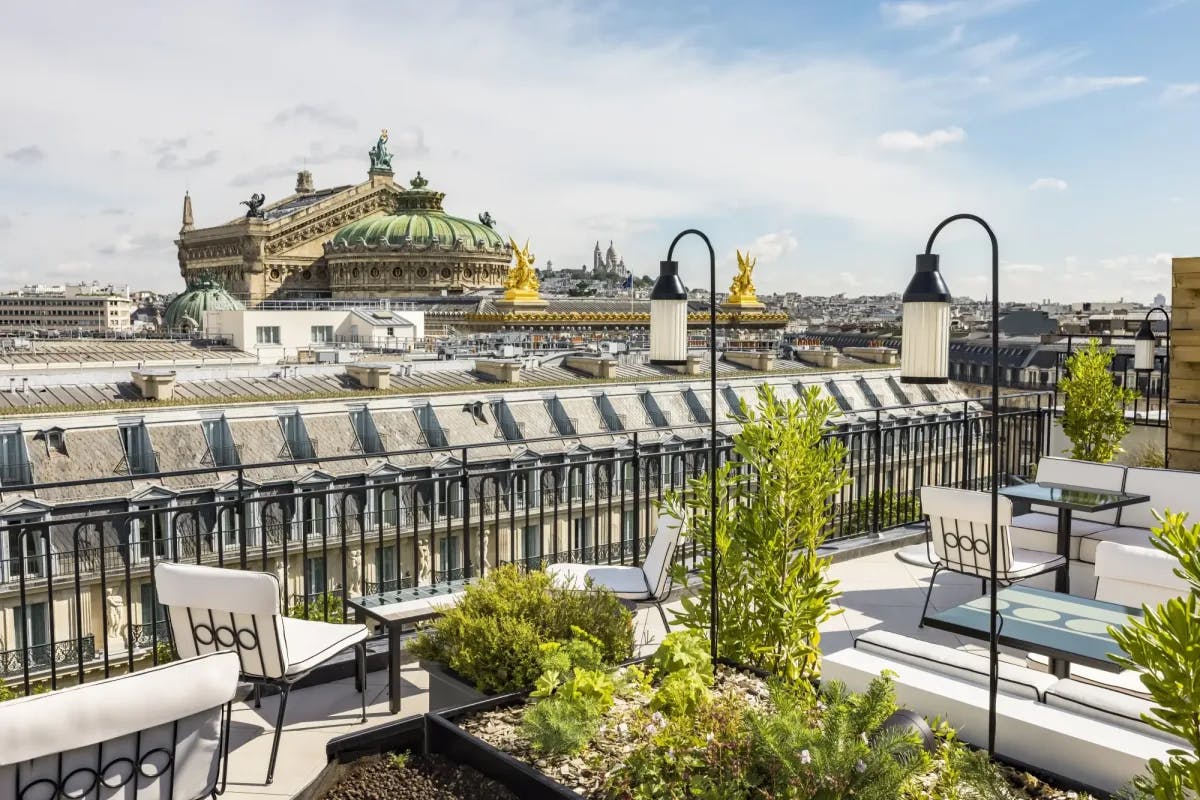 a fancy rooftop terrace filled with plants and tables and chairs overlooks a regal, ornate European building