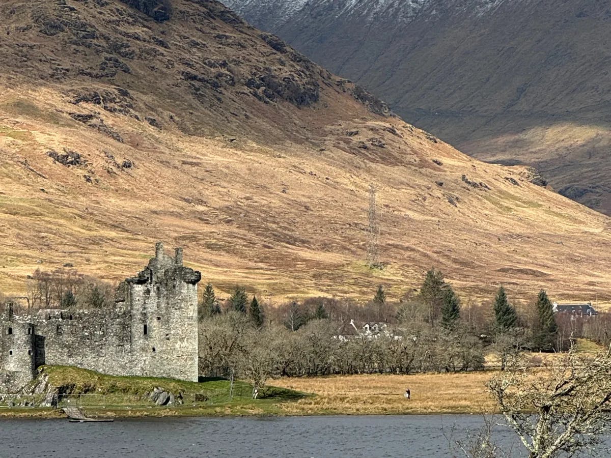 A historic stone castle by a tranquil water body, encircled by rugged mountains with snowy and barren slopes, exuding a sense of timeless beauty and natural splendor.