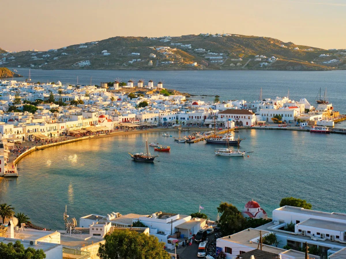 A panoramic view of the Mykonos harbor at sunset, showcasing white buildings against the Aegean Sea, with boats in the water and hills in the distance.