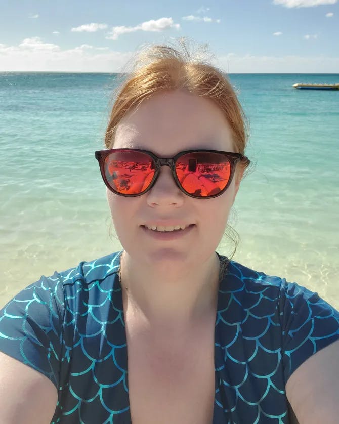 Picture of Jeanne at beach wearing a blue patterned top and orange tinted sunglasses