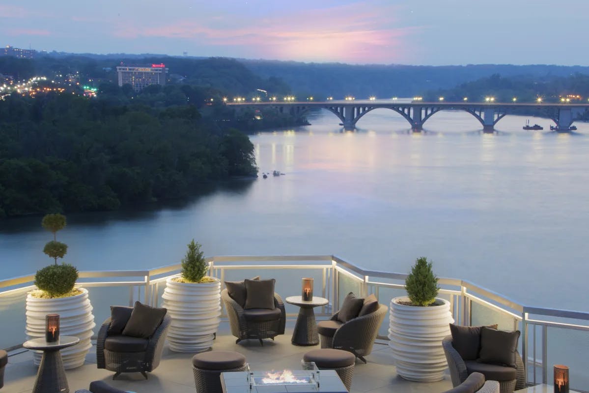 A view of the Potomac River and Key Bridge beyond a rooftop bar set with gray chairs and white planters.