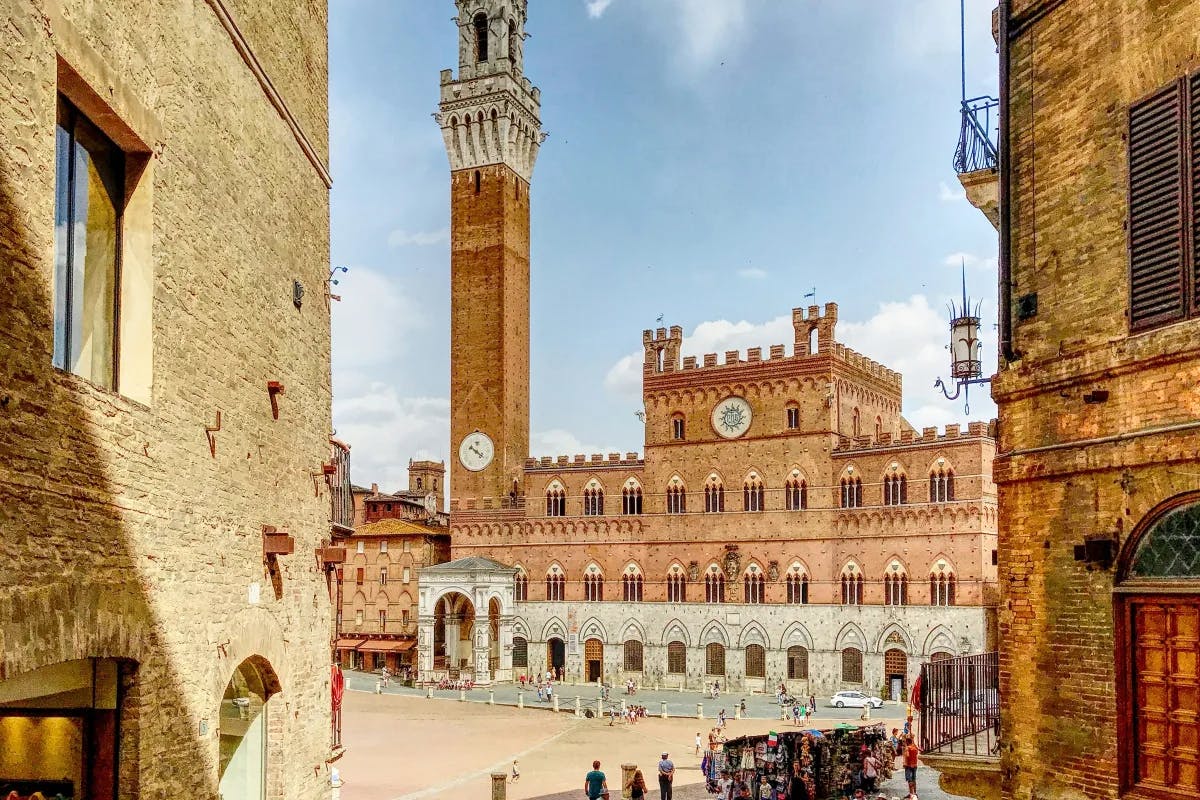 A beautiful stone square with elaborate buildings, a piazza, clock tower and people walking around in the distance. 