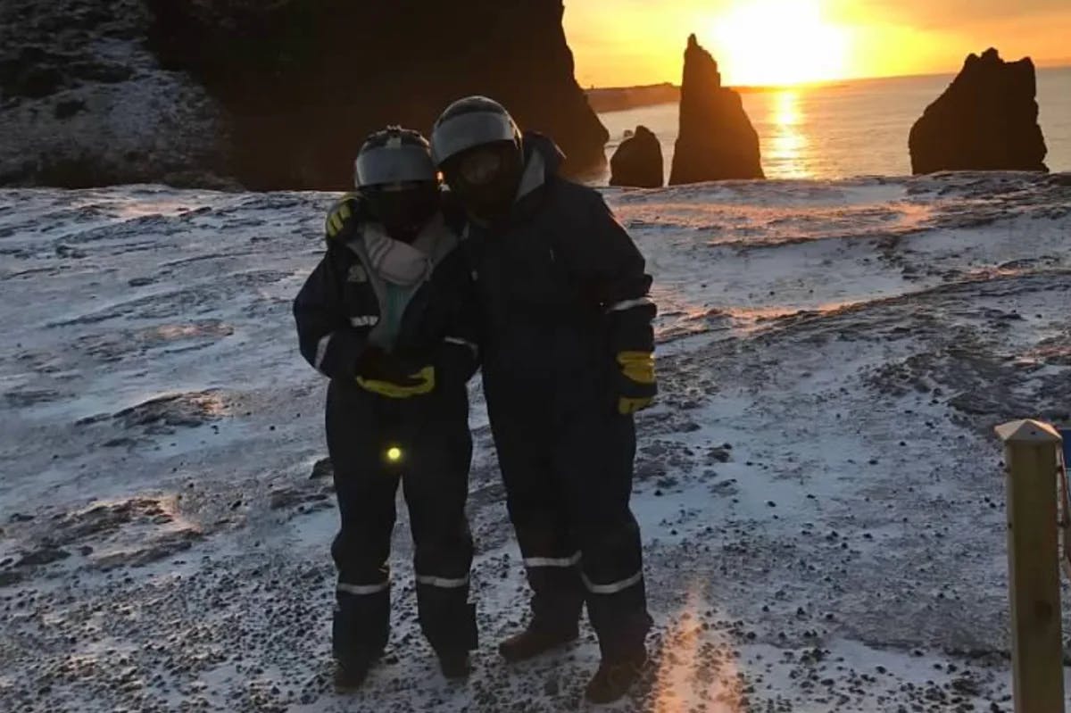 Two people dressed in outdoor gear preparing to snowmobile outside with snow, rocks and a vibrant orange sunset in the background. 