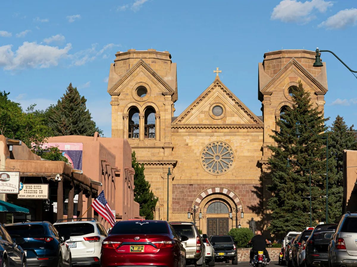 The image displays a historic church with twin bell towers and a rose window, nestled among trees and parked cars under a clear blue sky.