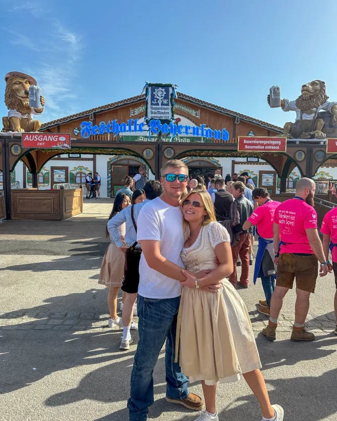 Hannah in a beige dress with her arm around a man in front of the entrance to an Oktoberfest event on a sunny day