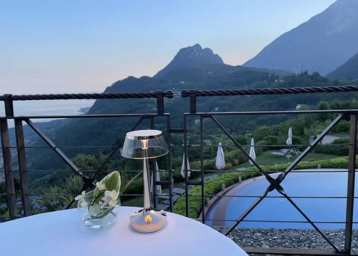 A view of a lamp on top of a white table top outside next to a balcony fence overlooking a swimming pool, mountains and grassy lawn. 