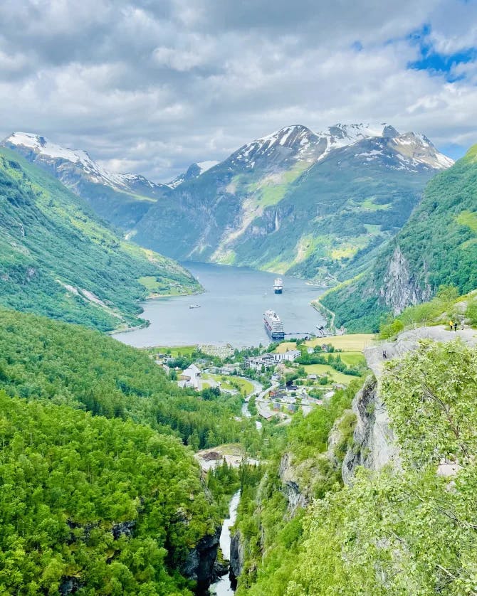 A beautiful view of Geirangerfjord with green trees, a body of water and snowy peaks in the distance.