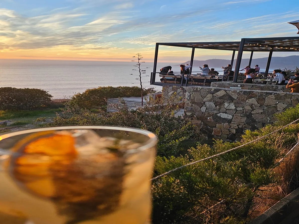 The image depicts a glass with a beverage in the foreground, set against a scenic backdrop of a pier, people, and the ocean under an evening sky.