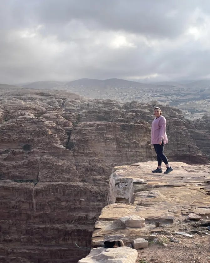 Kathryn wearing a pink top and black pants while standing on the edge of a rocky cliff with large rock formations in the background.