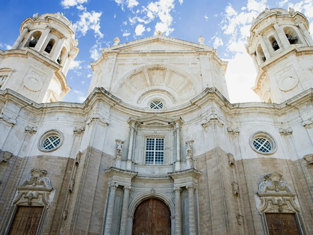 The image shows the facade of a grand, baroque-style cathedral with two bell towers under a blue sky with clouds.