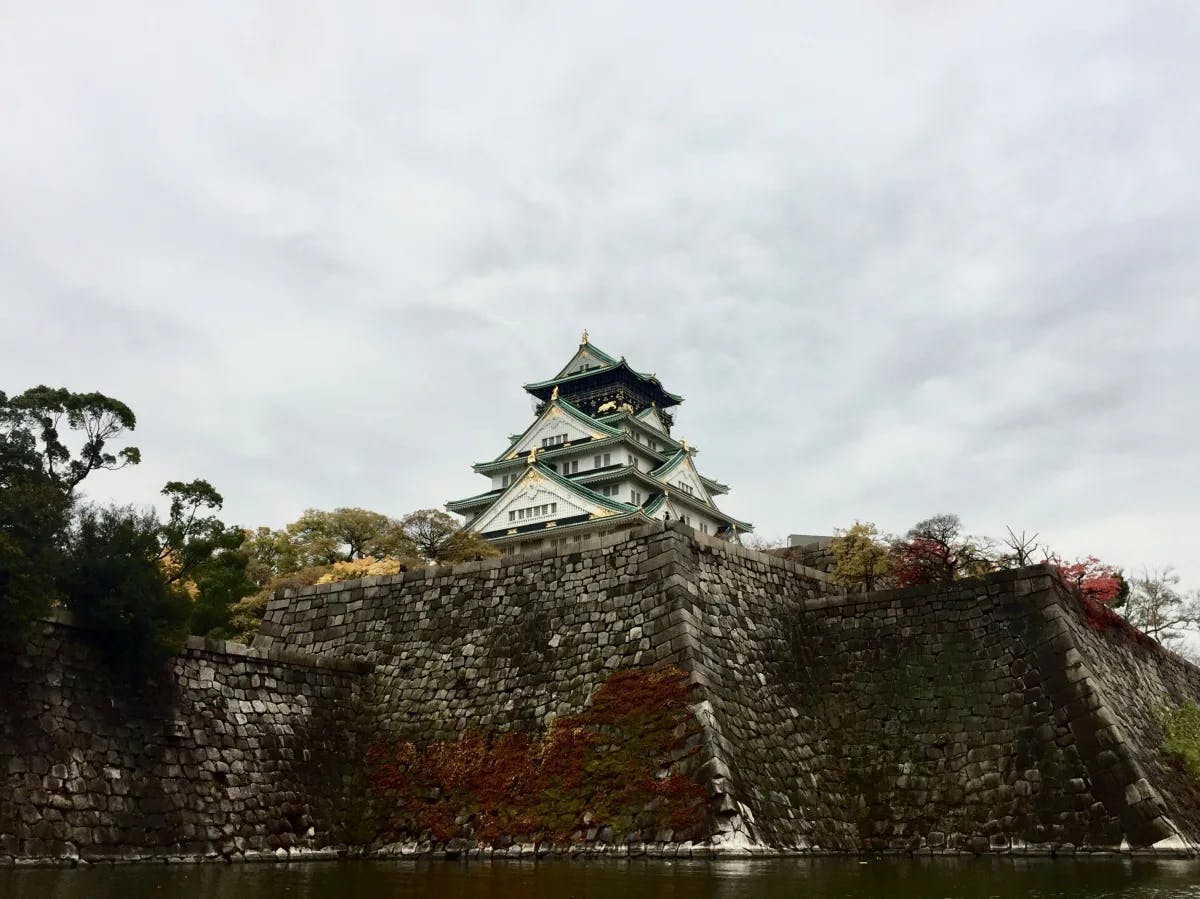 A traditional Japanese castle with multiple tiers and a surrounding stone wall is set against a cloudy sky.